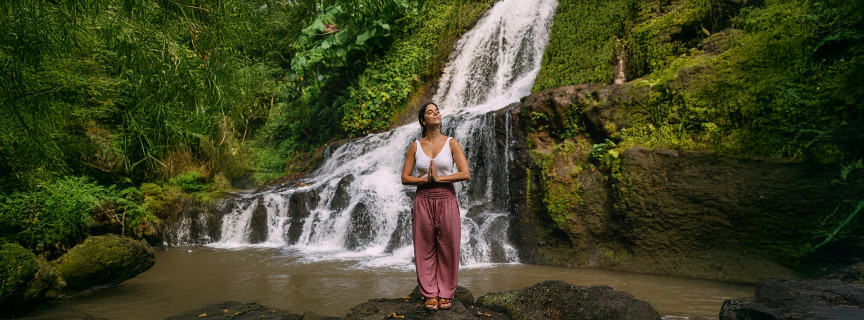Frau steht entspannt vor einem Wasserfall und genießt den Moment – gekleidet in einer bequemen Haremshose.