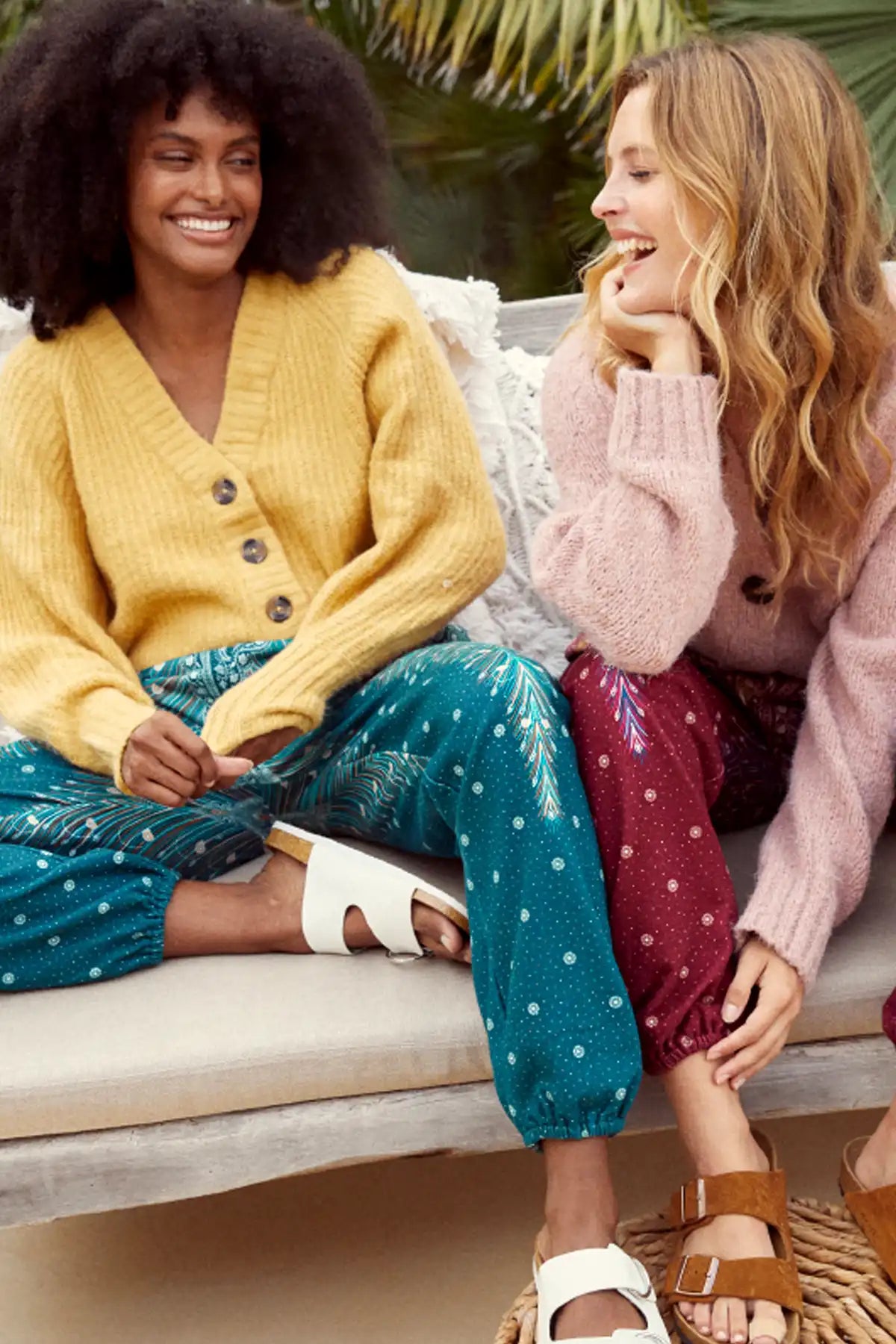 Eine lächelnde Frau kombiniert Haremshose in gemusterten Grüntönen mit gelber Strickjacke. Ihre Freundin kombiniert Haremshose in gemusterten Rottönen mit rosa Strickjacke.