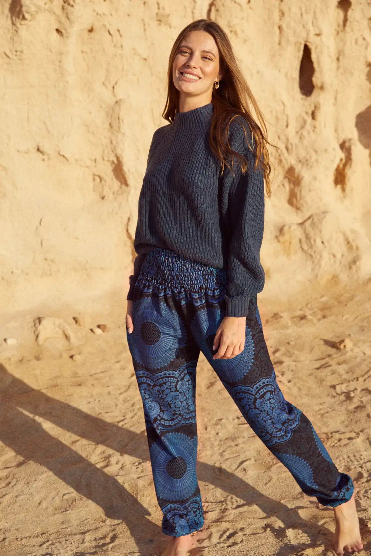 Frontalansicht einer stehenden Frau im Sand, die die gemusterte Haremshose in dunklen Blautönen mit einem dunkelblauen Strickpullover kombiniert.