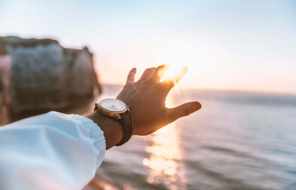 Person mit Armbanduhr streckt den Arm Richtung Horizont mit Sonnenuntergang am Meer