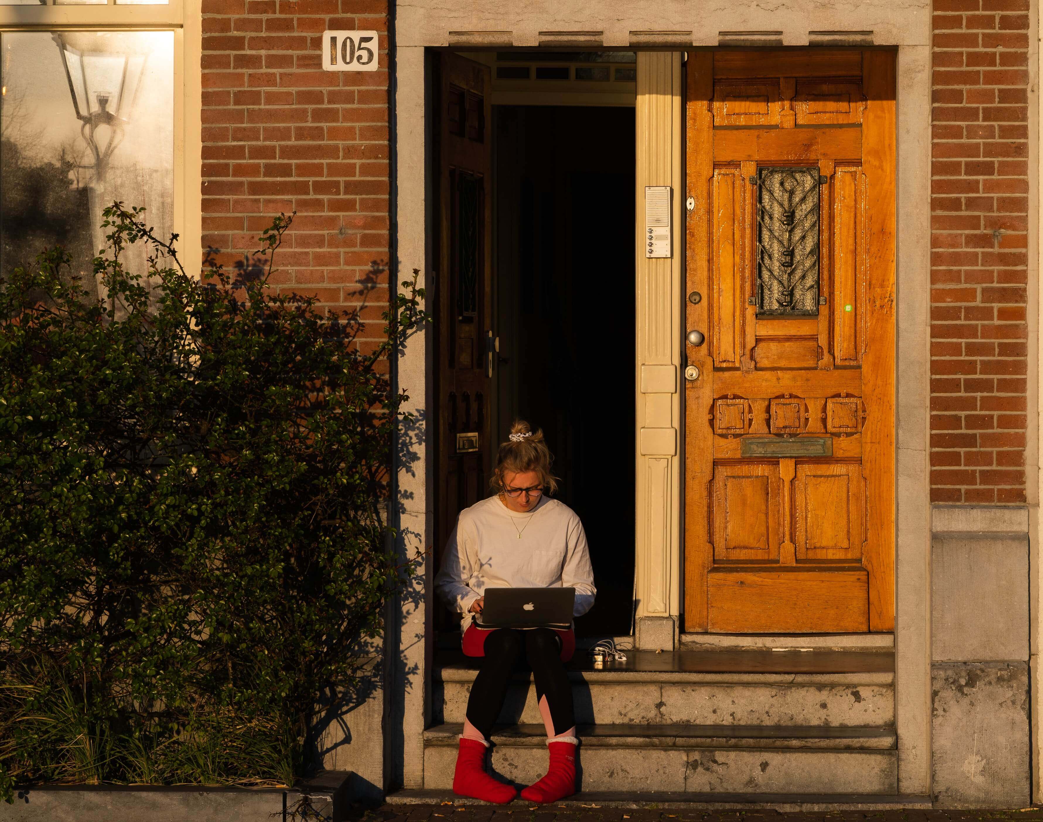 Person sitzt mit einem Laptop in der Sonne auf den Stufen vor einem Haus