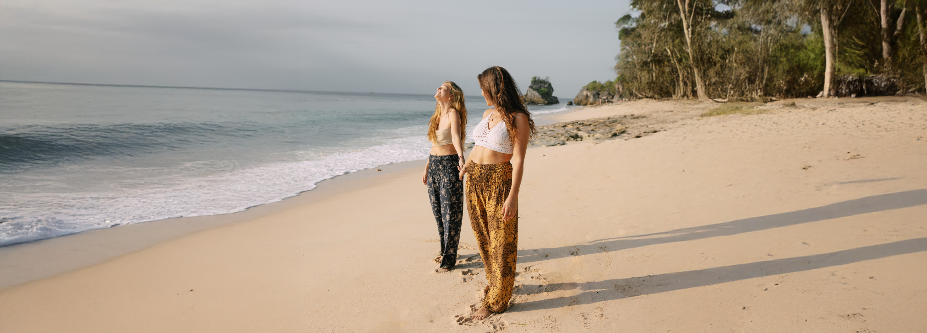 Zwei Frauen stehen Hand in Hand am Strand vor dem Meer mit sanften Wellen und wirken glücklich.