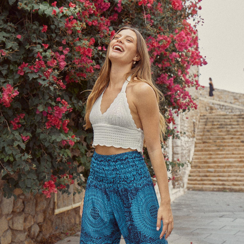 Lachende Frau in blauer Haremshose und weißem Top steht vor einer bunt bepflanzten Steinmauer